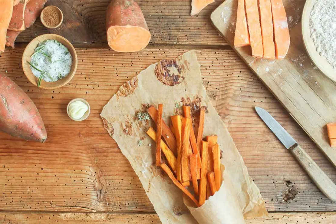 Pommes aus Suesskartoffeln liegen in einer Tuete auf einem Tisch mit Backpapier, Gewuerzen und Sauces. [Foto: AdobeStock_eflstudioart]