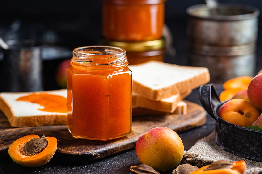 Das Bild zeigt ein Glas mit Aprikosenmarmelade, das auf einem rustikalen Holztisch steht. Im Hintergrund befinden sich zwei Scheiben Brot, eine davon bereits mit Marmelade bestrichen. Um das Glas herum liegen frische Aprikosen, einige davon aufgeschnitten, sodass der Stein sichtbar ist. [Foto: AdobeStock_elenavah]