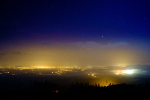 Eine nächtliche Landschaft zeigt Lichtverschmutzung über einer Stadt. Der Himmel ist tiefblau mit Sternen, während orangefarbene Lichter der Stadt die untere Hälfte des Bildes erhellen. Dunkle Hügel oder Wälder im Vordergrund kontrastieren mit dem künstlichen Licht. [Foto: AdobeStock_Miguel]