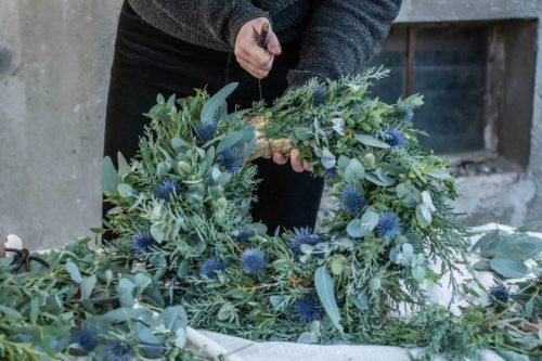 Eine Frau bindet einen Adventskranz aus mit Eukalyptus, Distel, Mimose und Zypresse und zieht das Gesteck mit Draht fest.