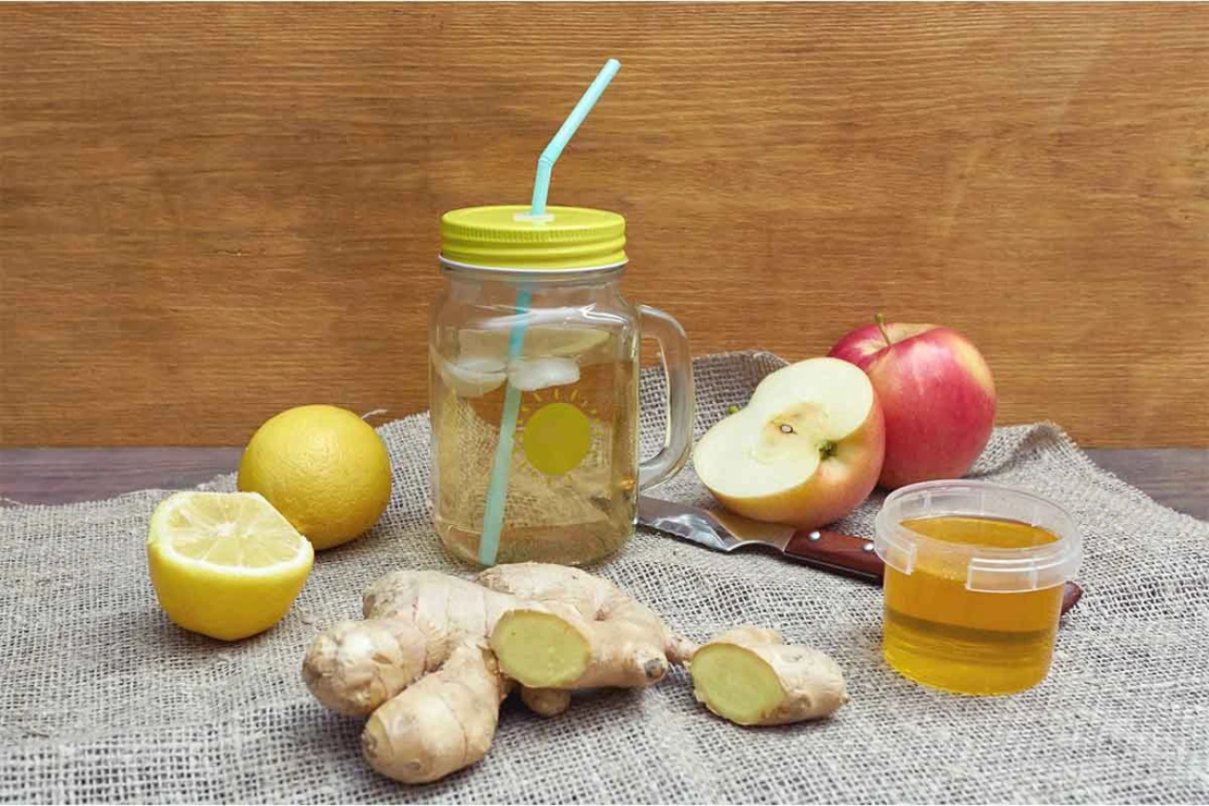 Swizzle oder auch Switchel in einem Mason-Jar mit gelbem Deckel und Strohhalm, der mit einem aufgeschnittenen Apfel, einem angeschnittenen Stueck Ingwer, Honig und einer Zitrone auf einem Jutetuch arrangiert ist. [Foto: AdobeStock_205237292_Alrandir]